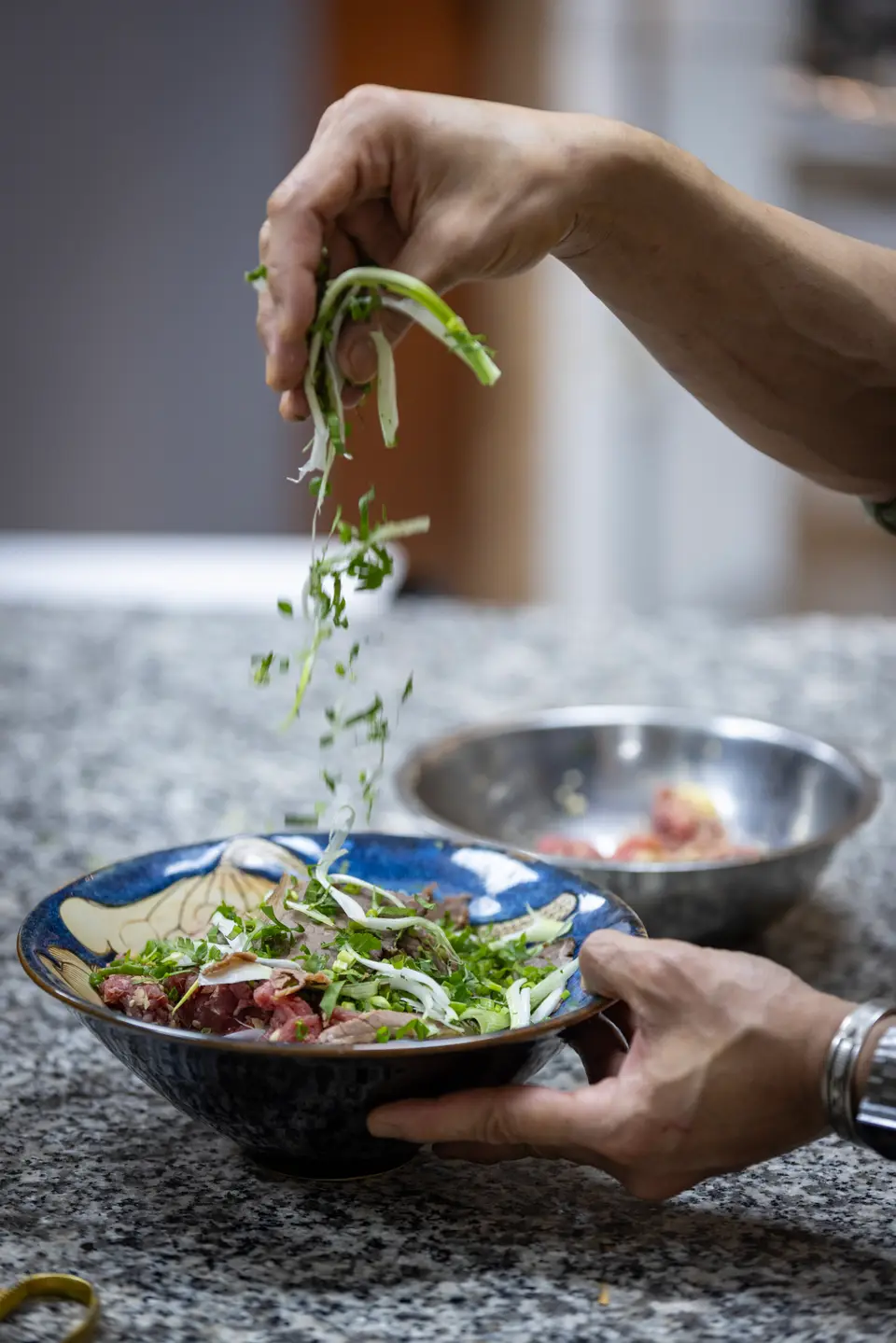 Des mains parsèment un bol de phở d'herbes fraîches ciselées, dans la cuisine du Chợ Vỉa Hè.