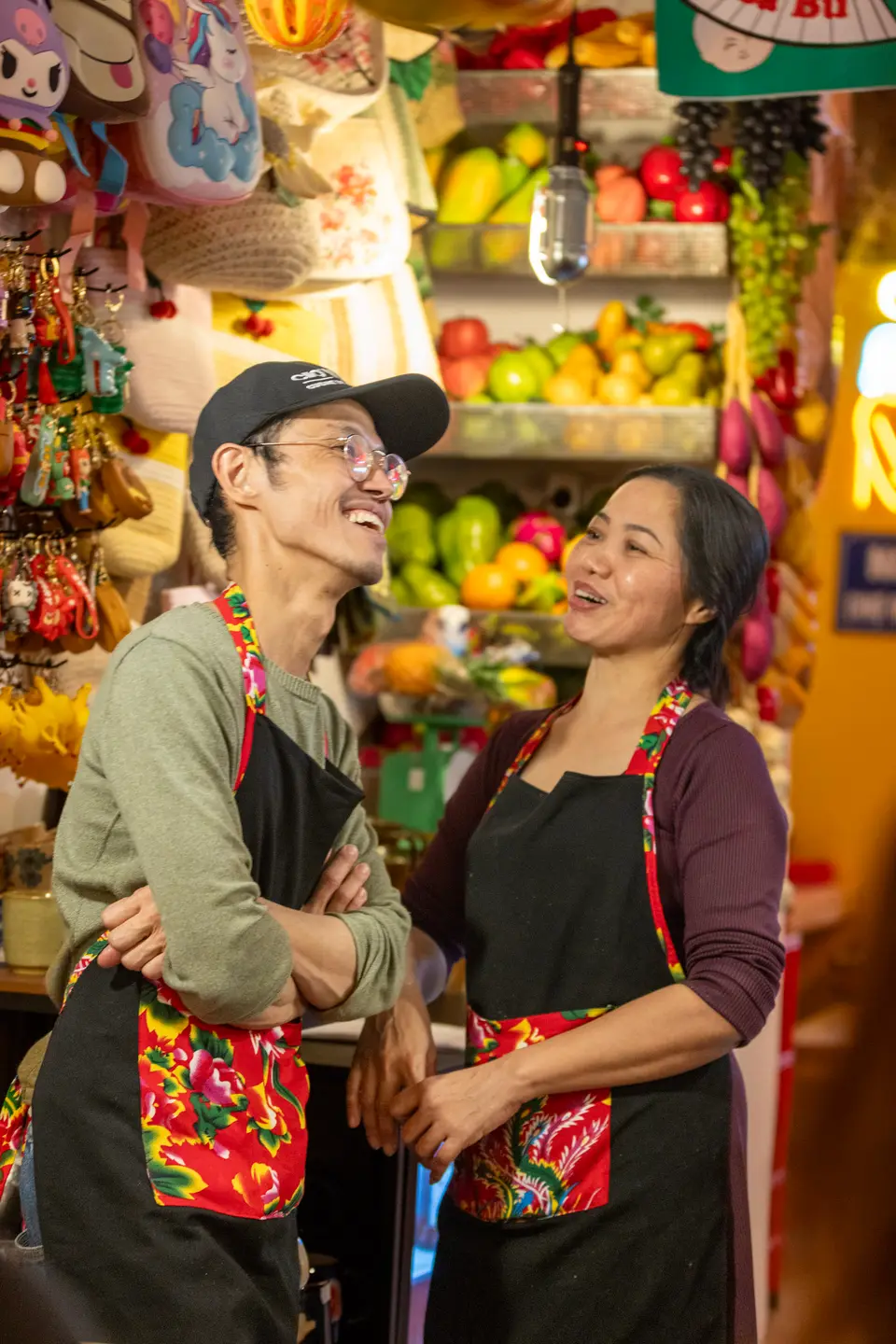 Le couple fondateur du Chợ Vỉa Hè, en tabliers fleuris, riant devant le stand aux couleurs du marché.